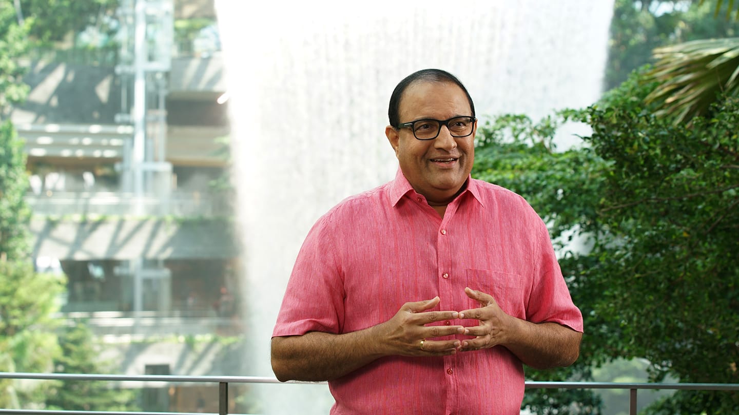 Man in pink shirt and glasses gesturing in front of a waterfall and greenery.